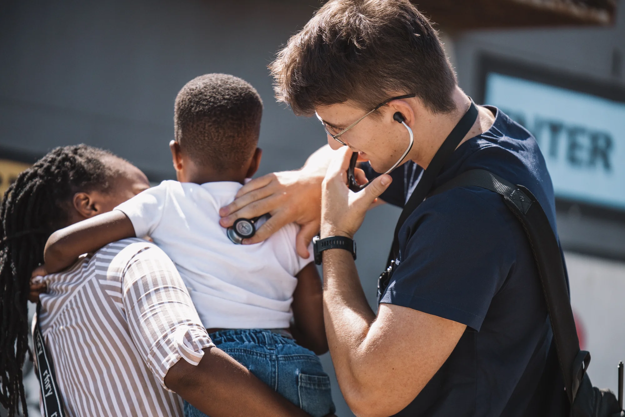 A doctor listens to a child's heartbeat with a stethoscope on a sunny street.
