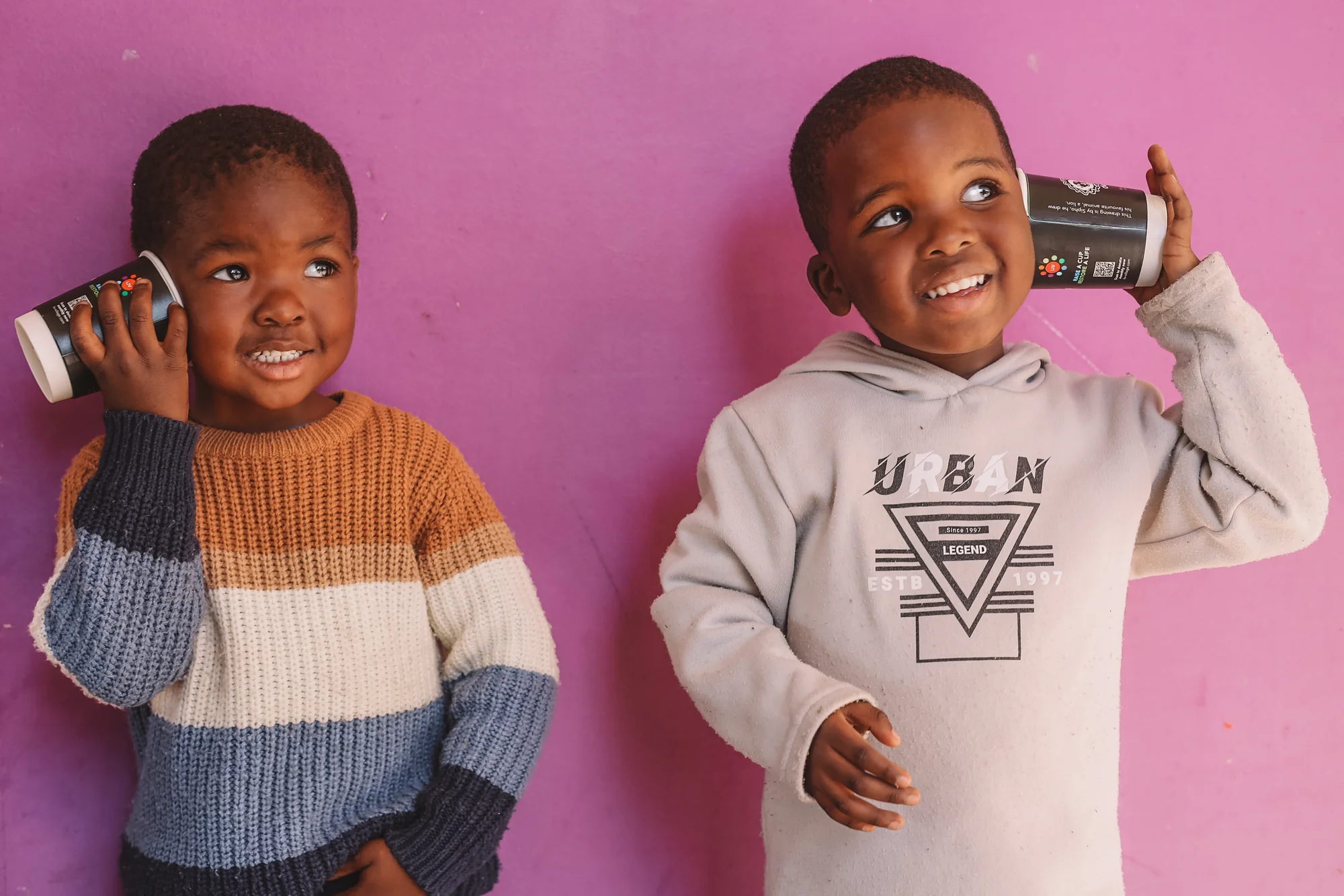 Two children with mugs, playing with a camera, pink wall