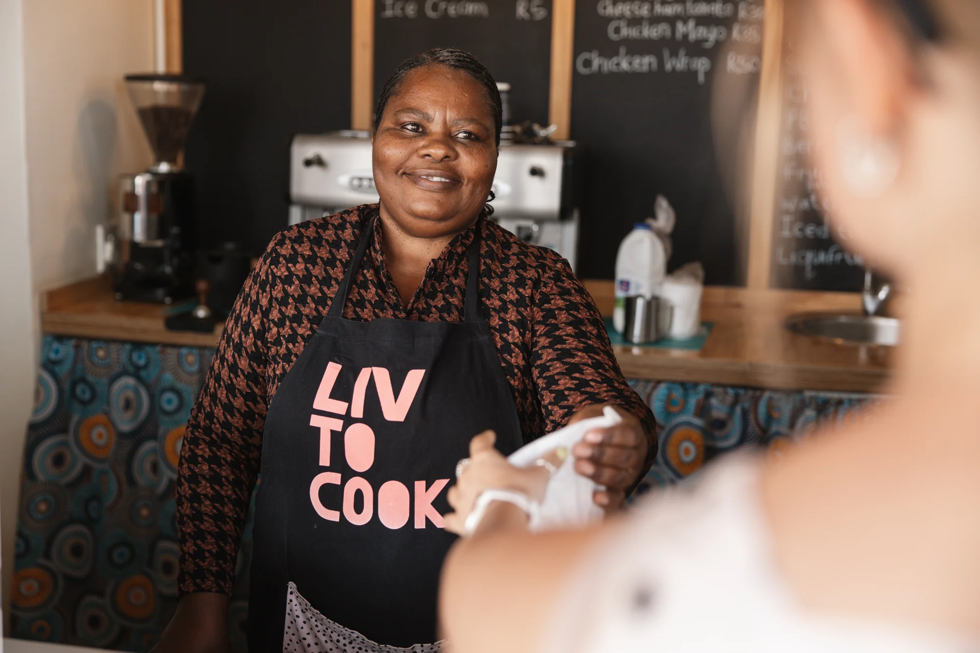 A woman at the coffee machine, 'Liv to Cook' apron