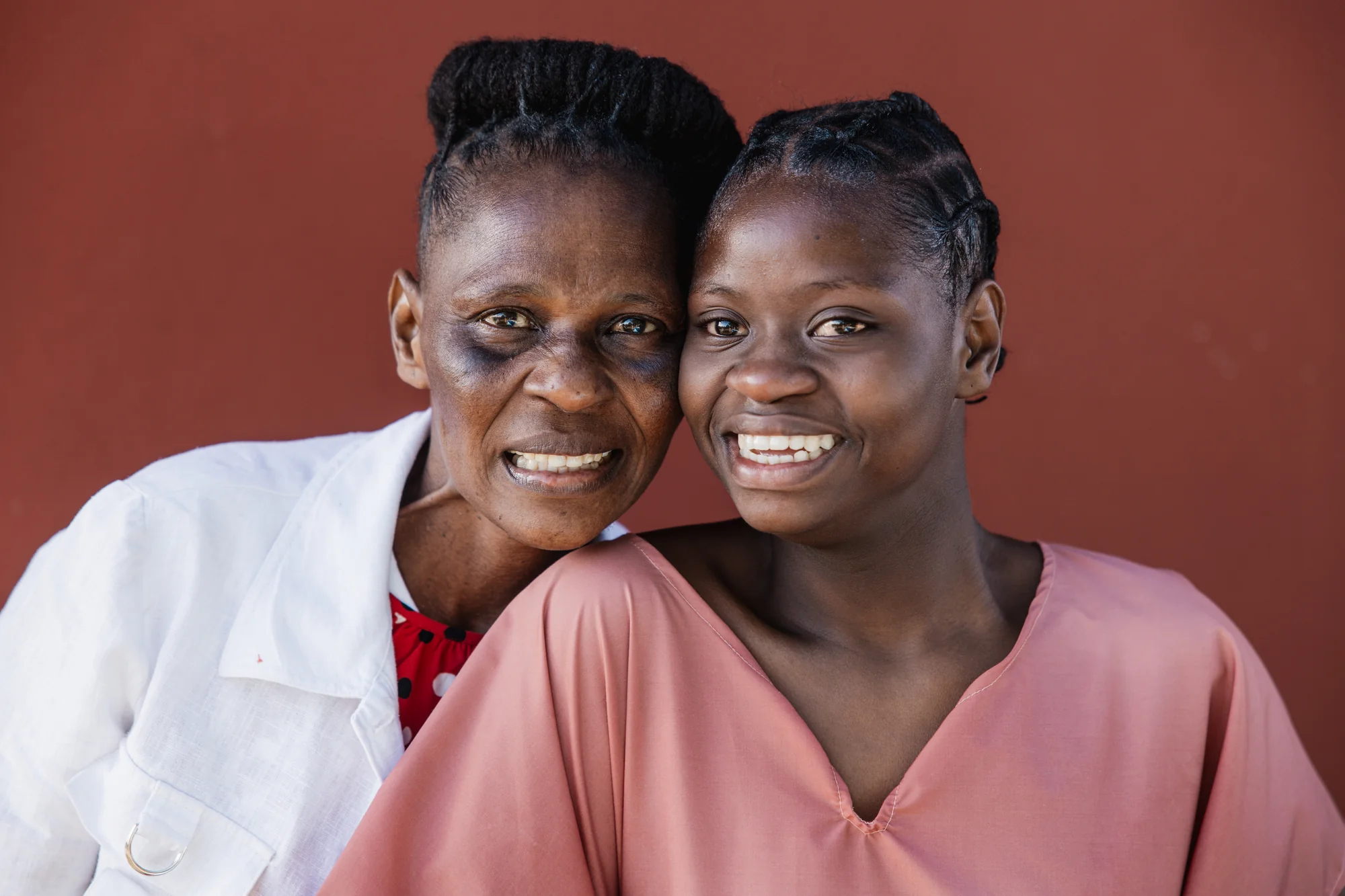 A mother and daughter smile cheek to cheek against a terracotta wall.