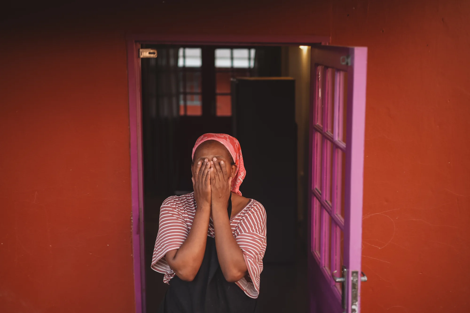 Mom Yvette laughs, covering her face with both hands, standing in a bright pink doorway.